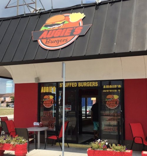 Exterior of Augie's Burgers, featuring a sign with a burger and fries, outdoor seating, and a bright red and black color scheme.