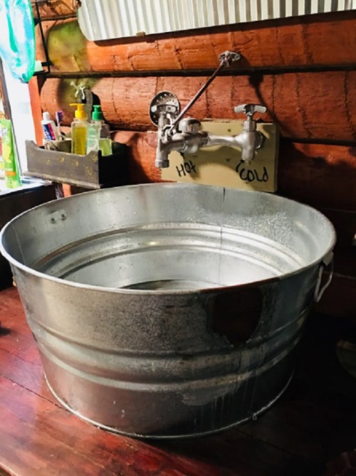 A large metal wash basin with hot and cold water taps, set on a wooden countertop in a rustic environment.