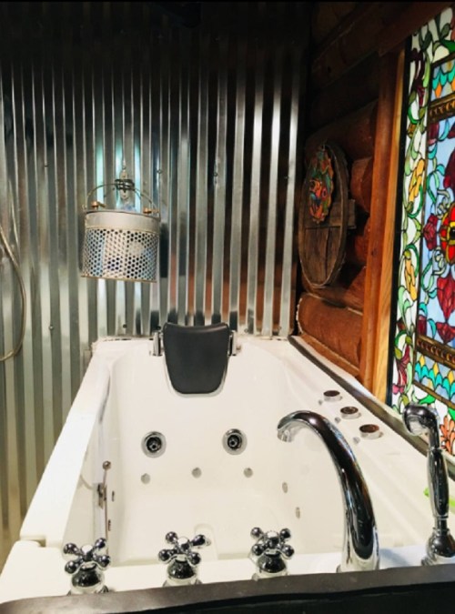 A modern white bathtub with chrome fixtures, set against a rustic metal wall and colorful stained glass window.