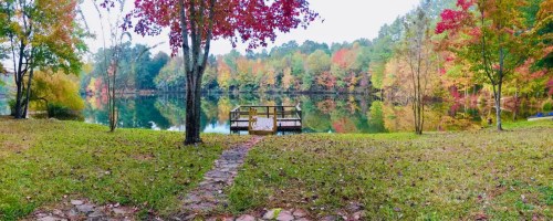 A serene lake view with colorful autumn trees and a wooden dock, surrounded by lush green grass and a stone path.