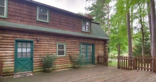 A rustic wooden cabin with green doors, surrounded by trees and a wooden deck.