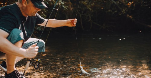 A man crouches by a stream, holding a fishing rod with a caught fish just above the water's surface.
