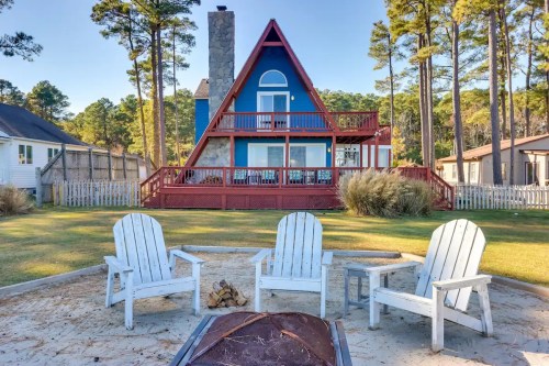 A-frame house with a stone chimney, surrounded by trees, viewed from a fire pit area with white chairs in the foreground.