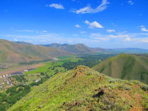 A panoramic view of rolling green hills and mountains under a bright blue sky with scattered clouds.