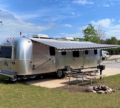 A shiny silver Airstream trailer parked on a concrete pad with a picnic table and fire pit nearby, under a blue sky.