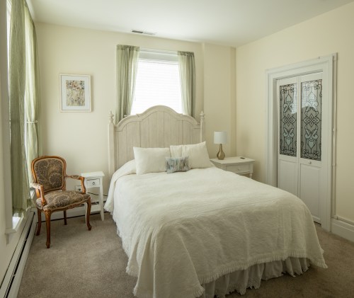 A cozy bedroom featuring a white bed, decorative chair, nightstand, and soft natural light from the window.