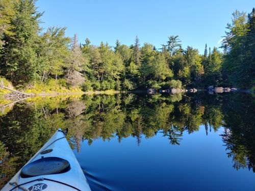 A serene lake scene with a kayak in the foreground, surrounded by lush trees and clear blue skies reflecting on the water.