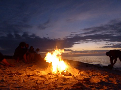 A beach bonfire at sunset, with silhouettes of people sitting nearby and a calm sea in the background.