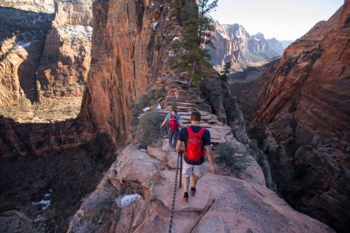 Two hikers with red backpacks walk along a narrow rocky path in a stunning canyon landscape.