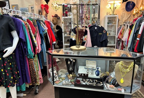 A vibrant thrift store interior filled with colorful clothing racks and a display case of vintage jewelry and accessories.