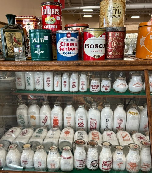 A display of vintage coffee cans and glass milk bottles on a wooden shelf in an antique store.