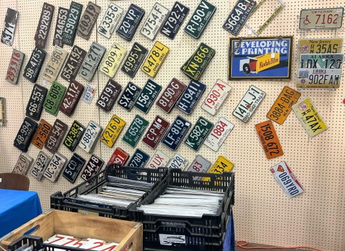 A wall display of various vintage license plates, with crates of more plates in the foreground.