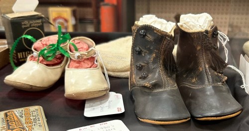 Two pairs of vintage children's shoes displayed in a glass case, one white and one black, with decorative details.