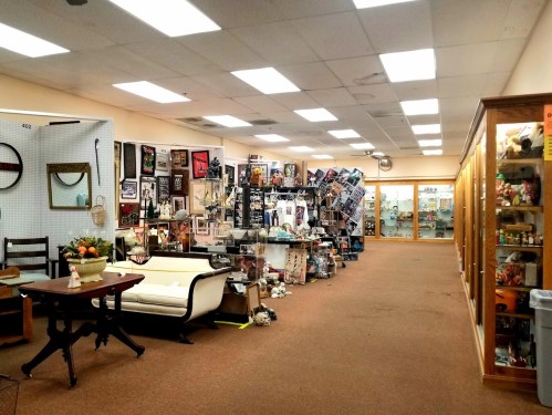A spacious antique shop with various collectibles, furniture, and display cases lining the walls. Brightly lit interior.