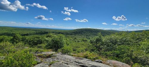 A panoramic view of lush green hills under a bright blue sky with scattered clouds. Rocky foreground adds texture.