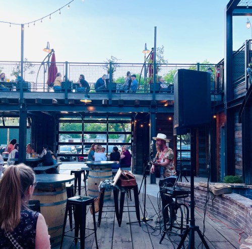 A musician performs on stage at an outdoor venue with patrons seated at tables, enjoying the evening atmosphere.