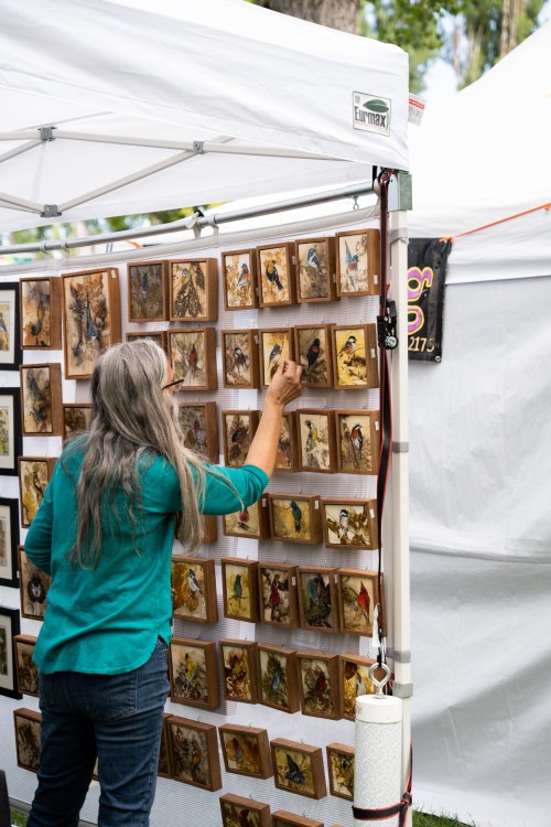 A woman examines artwork displayed on a wall at an outdoor art fair under a white tent.