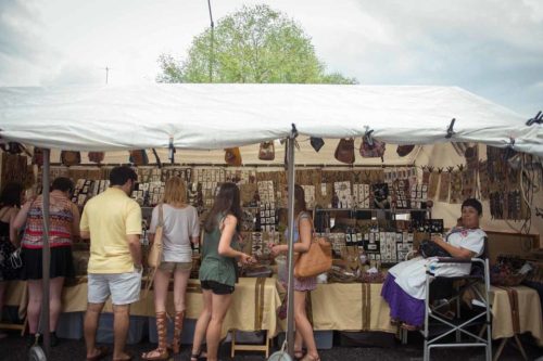 A market stall with various handmade crafts, people browsing, and a woman sitting nearby in a wheelchair.
