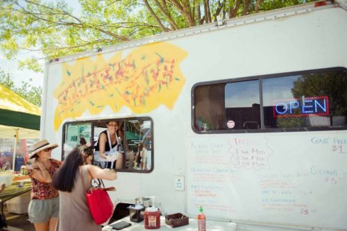 A food truck with a colorful sign, serving customers at a market, with a menu displayed on the side.