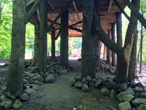 A pathway lined with rocks leads under a wooden structure supported by tree trunks, surrounded by greenery.