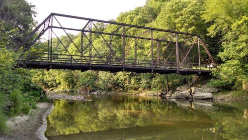 A rusty metal bridge spans a calm river, surrounded by lush green trees and rocky banks under a clear sky.