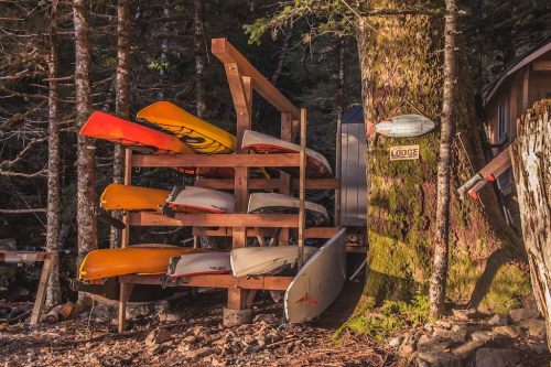 A wooden rack holds colorful kayaks beside a tree, with a sign reading "Lodge" in a forested area.