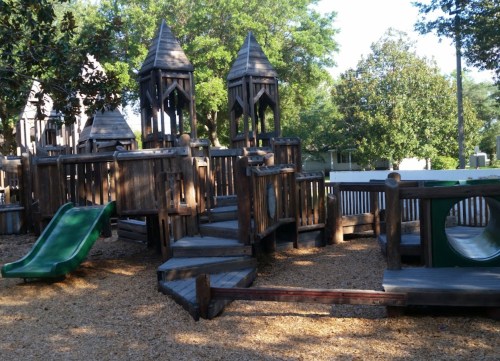 Wooden playground structure with slides, towers, and climbing areas, surrounded by trees and wood chips.