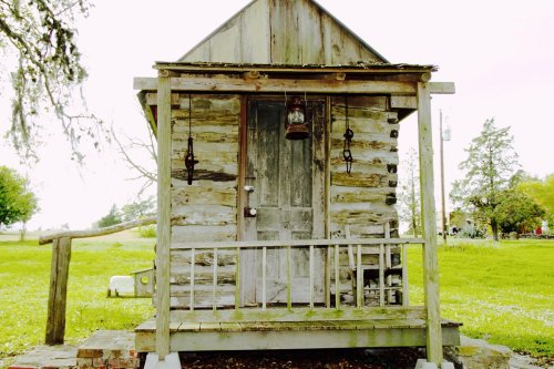 A rustic wooden cabin with a porch, featuring a weathered door and lanterns, set in a grassy landscape.