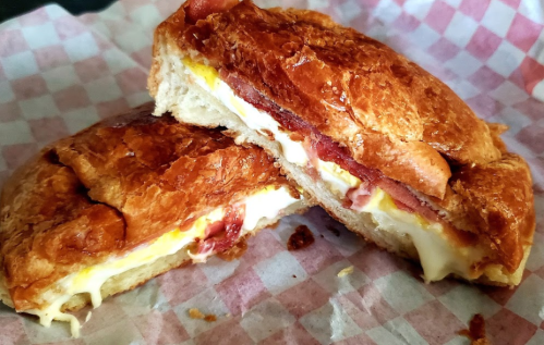 A close-up of a toasted croissant sandwich filled with melted cheese and ham, resting on a checkered paper.