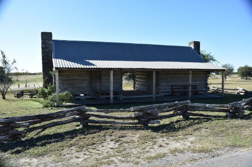 A rustic log cabin with a metal roof, surrounded by a wooden fence and open fields under a clear blue sky.
