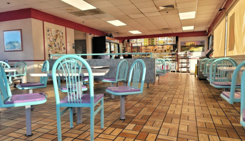 A brightly lit fast-food restaurant interior with colorful chairs and tables, featuring a tiled floor and menu board in the background.