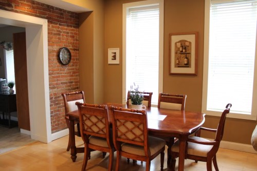 A cozy dining area featuring a wooden table, chairs, and a brick wall with framed art and a clock.