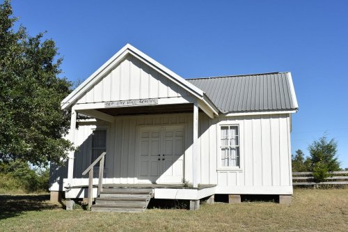 A small, white wooden building with a metal roof and steps, set against a clear blue sky and grassy area.