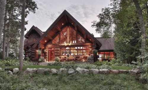 A large wooden cabin surrounded by trees, featuring a stone pathway and large windows reflecting warm light.