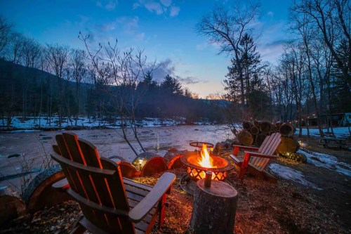 A cozy campfire by a frozen river at dusk, with wooden chairs and trees in the background.