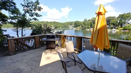 A sunny lakeside deck with a yellow umbrella, a grill, and a table overlooking calm water and trees.