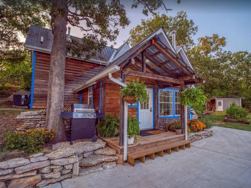 A charming wooden house with a porch, grill, and hanging plants, surrounded by greenery and colorful flowers.