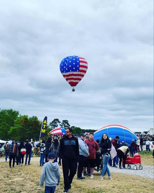 A crowd at a festival with a large American flag-themed hot air balloon overhead and colorful balloons in the background.