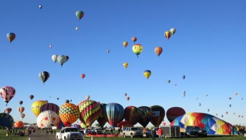 A vibrant sky filled with colorful hot air balloons, with a crowd and vehicles on the ground below.