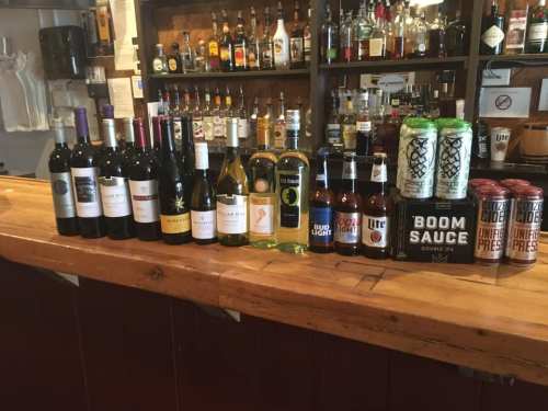 A variety of wine and beer bottles arranged on a wooden bar, with a selection of spirits in the background.