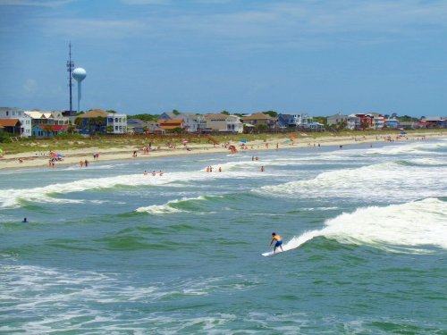 A surfer rides a wave near a beach filled with people, with colorful houses and a tower in the background under a blue sky.
