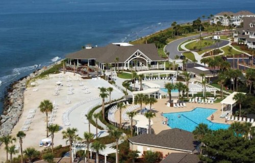 Aerial view of a coastal resort with a pool, lounge chairs, and palm trees along the shoreline.