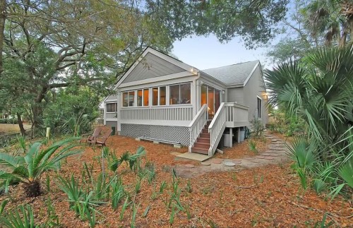 A cozy gray house surrounded by greenery and mulch, featuring a porch and steps leading to the entrance.