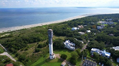 Aerial view of a coastal area featuring a tall lighthouse, sandy beach, and lush greenery with nearby houses.