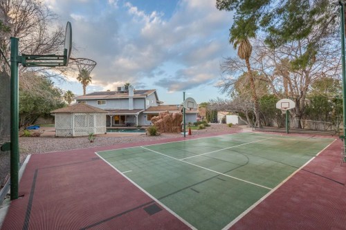 Outdoor basketball court with two hoops, surrounded by trees and a house in the background under a cloudy sky.