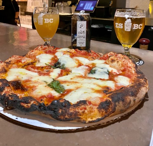 A close-up of a pizza with melted cheese and basil, flanked by two glasses of beer and a can on a bar counter.