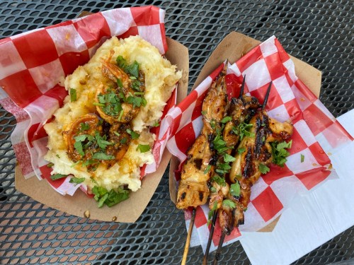 Two food trays on a black mesh table: one with shrimp over mashed potatoes, the other with grilled chicken skewers.