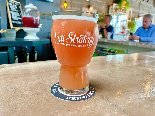 A glass of beer with a logo from Exit Strategy Brewing Co. sits on a bar counter with a blurred background.