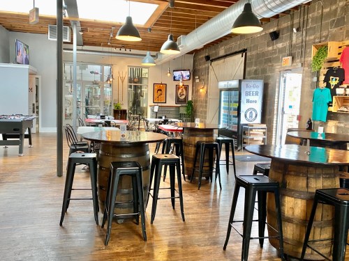 A spacious bar area with wooden tables, stools, and large windows, featuring a casual, inviting atmosphere.