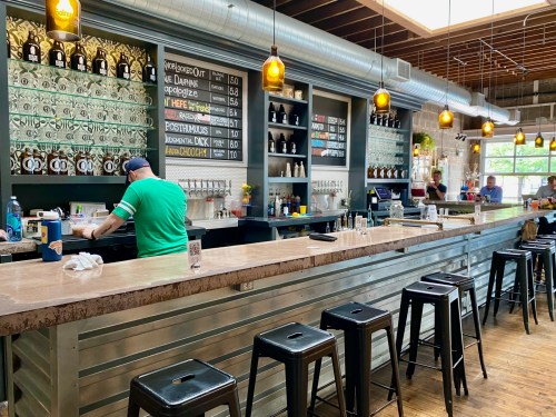 A modern bar with a metal counter, stools, and a chalkboard menu; staff are preparing drinks in a lively atmosphere.